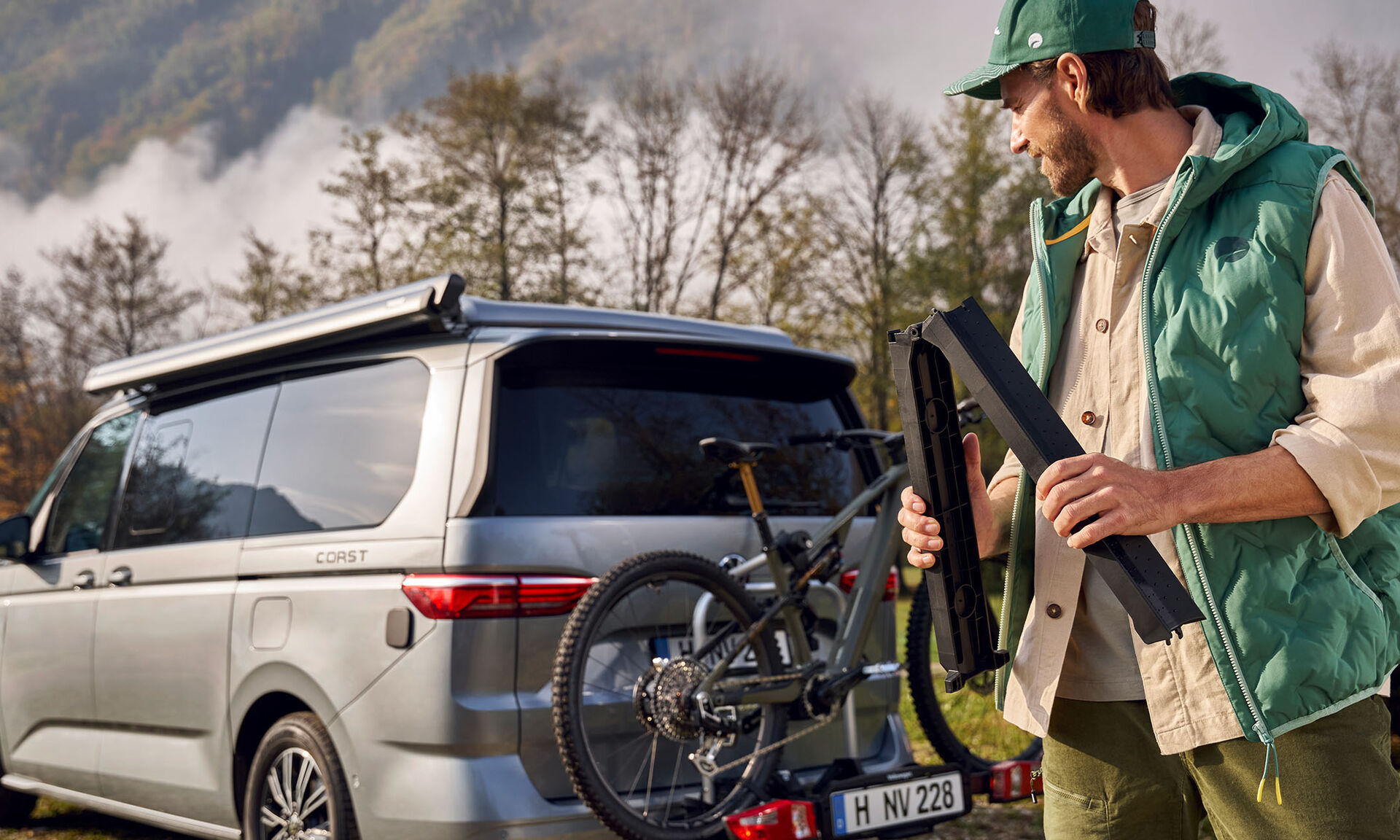 Držák na jízdní kolo na VW T7 a muž v zelené vestě California a zelené kšiltovce California na hlavě